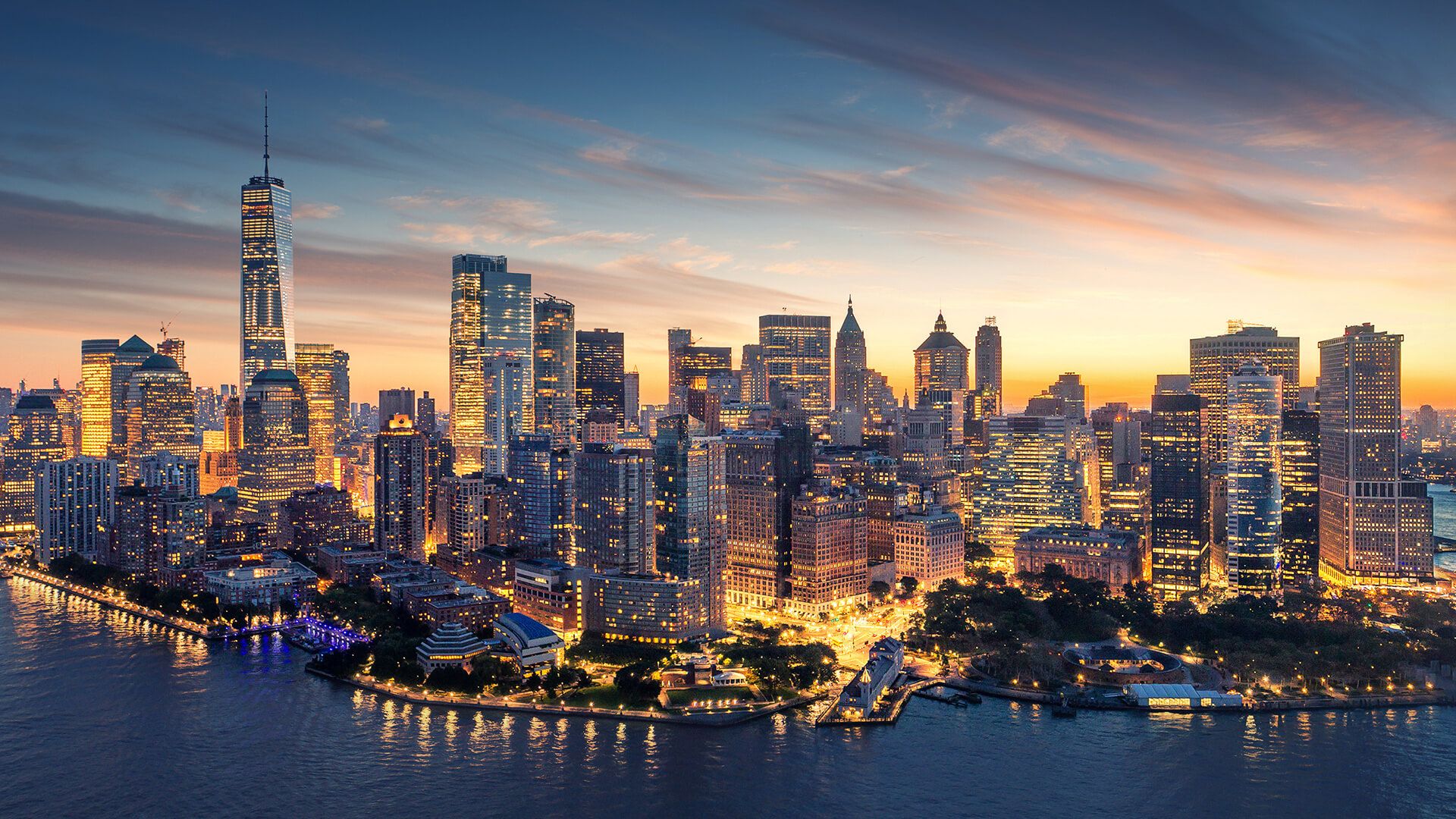 Aerial view of New York City skyline at sunset, showcasing illuminated skyscrapers and waterfront reflections.